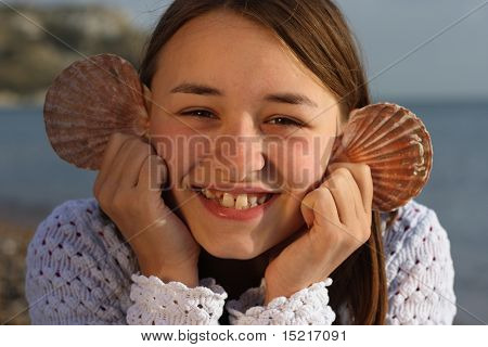 Young girl by the sea with seashells for ears smiling.