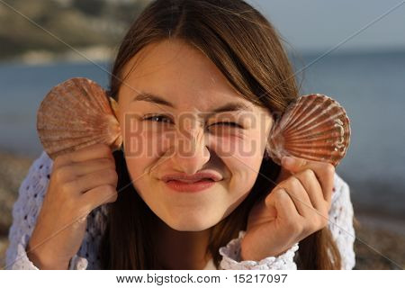 Young girl by the sea with seashells for ears pulling a face.