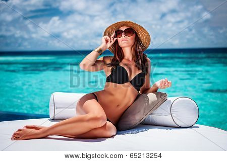 Sexy woman sitting on cozy white lounger on the beach, stylish model wearing fashionable hat and sunglasses, luxury summer vacation concept 