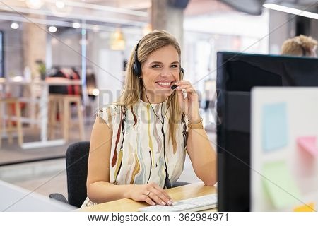 Smiling mid woman working as customer support operator with headset in a call center. Portrait of happy sales agent sitting at desk and looking at camera. Customer care support service representative.