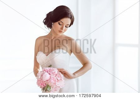 Beautiful bride perfect style. Wedding hairstyle make-up luxury wedding dress and bride's bouquet. Young attractive multi-racial Asian Caucasian model like a bride against white room at studio looking at bouquet.