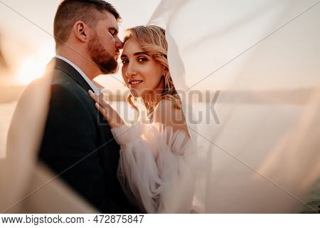 The Bride And Groom Under The Veil On The Sunset By The River