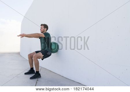 Strength training man doing squats using medicine ball rolling on wall squatting at fitness centre. Workout squat bodyweight exercises using medicine ball. Fitness athlete working out.