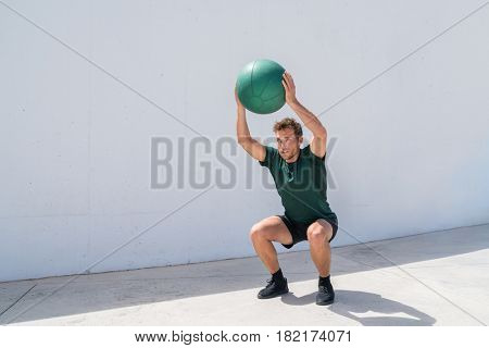 Squat with overhead medicine ball throw or shoulder press. Weight exercises workout fitness man training legs and glutes with weighted medicine ball squats. Athlete working out at gym.