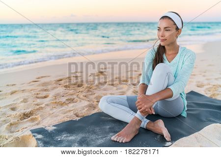 Yoga beach woman relaxing on fitness mat after workout. Fit Asian fitness athlete girl doing exercise in activewear at sunset or sunrise. Mindfulness healthy lifestyle.