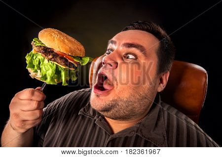 Diet failure of fat man eating fast food hamberger. Happy overweight person with wide-open mouth who spoiled healthy food by greedily eating huge hamburger on fork. Junk meal leads to obesity.