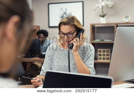 Mature businesswoman talking on phone in office. Casual business woman talking on landline phone in a creative agency. Beautiful blonde receptionist with eyeglasses taking client details over phone.