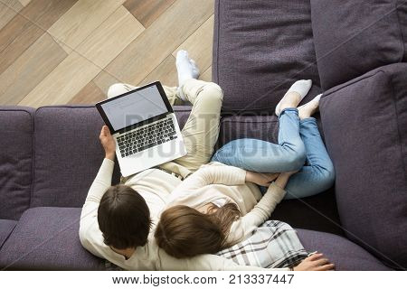 Young loving couple sitting on comfortable couch embracing holding laptop at home, man and woman hugging relaxing on sofa in cozy living room using computer browsing web sending e-mails, top view