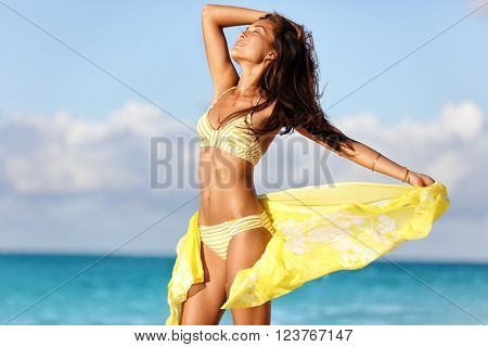 Sexy suntan woman relaxing enjoying sunset on beach with beachwear cover-up wrap showing slim bikini body for weight loss and skin care epilation concept. Asian model sunbathing during summer holiday.