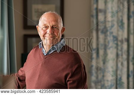 Portrait of happy retired senior man standing at home near window. Satisfied old man looking at camera and smiling while standing near the window. Positive and confident elderly enjoy his retirement.