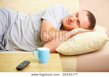 Retrato de un hombre durmiendo en el sofá en casa con almohadas bajo la cabeza. Taza azul y TV controller para