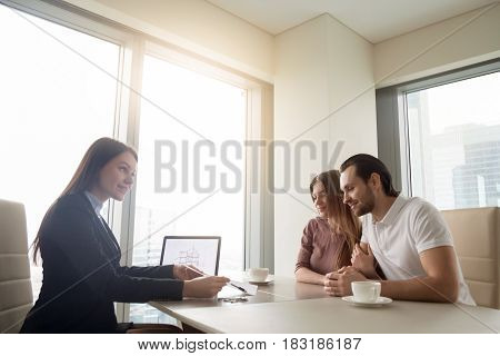 Female realtor meeting with young couple in real estate agency office. Discussing first property investment with broker, construction company manager showing build project, apartment plan on a laptop