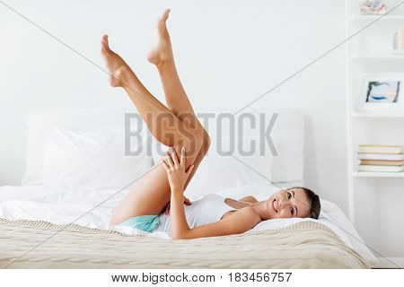 people, beauty, depilation, epilation and bodycare concept - beautiful woman lying on bed and touching smooth leg skin at home bedroom