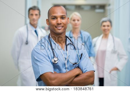 Confident male nurse in front of his medical team looking at camera. Smiling african young surgeon standing in front of his colleagues with folded arms. Multiethnic doctor with staff in background.