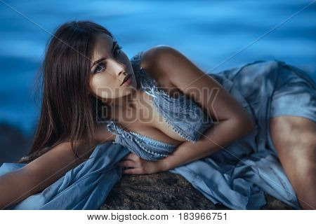 Fashion Art Photo. Model Girl Portrait in Moonlight at Night. Sexy Glamour Mystical Beautiful Woman in blue dress laying on rock over water background. Beautiful fashion creative shot of seductive multi-racial Asian Caucasian sexy female.