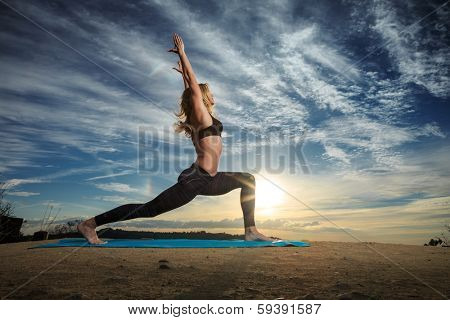 Woman practicing Warrior yoga pose outdoors over sunset sky background. 