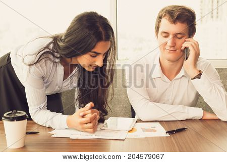 Indian assistant proving report to boss and waiting when he stopping talking on phone. Smiling young businessman listening to partner and looking through paper. Busy day concept