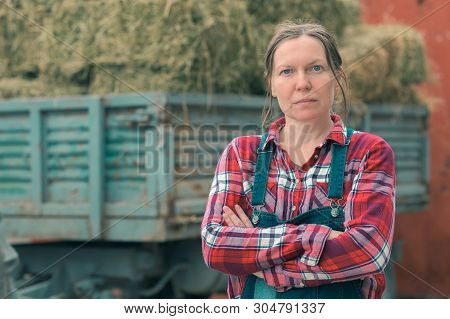 Female Farmer Posing In Front Of Hay Wagon. Portrait Of Woman Farm Worker In Plaid Shirt And Bib Ove