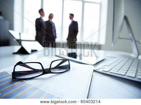 Technological devices, eyeglasses and financial document at workplace on background of three businessmen discussing ideas