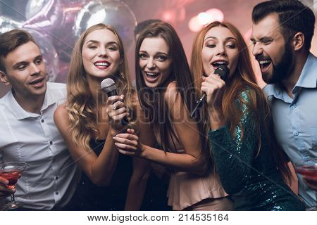 Three beautiful girls sing in a karaoke club. Behind them are men waiting for their turn. Young people have fun in a nightclub.