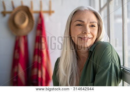 Portrait of beautiful senior woman sitting near big window at home and contemplate. Closeup face of fashionable old woman at home relaxing in living room. Mature woman thinking while looking away.