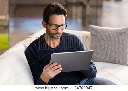 Man using digital tablet on sofa at home. Portrait of a serious man wearing eyeglasses working on digital tablet at home. Handsome man sitting on the sofa using tablet pc at home in the living room. 
