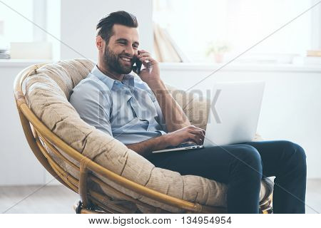 Working at home. Cheerful young handsome man working on laptop and talking on the mobile phone while sitting in big comfortable chair at home