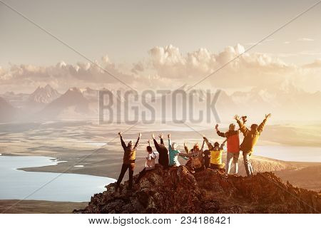 Big Group Of People Having Fun In Success Pose With Raised Arms On Mountain Top Against Sunset Lakes