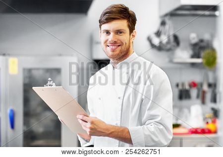 cooking, profession and people concept - happy smiling male chef cook writing to clipboard and doing inventory at restaurant kitchen