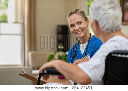 Friendly doctor examining health of patient sitting in wheelchair. Happy smiling nurse consulting disabled patient about treatment. Nurse caring about elder handicap man at home.