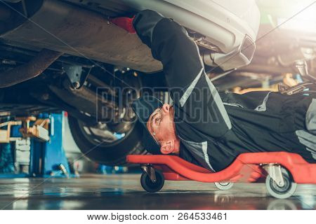 Caucasian Car Mechanic In His 30s Under The Car On The Mechanics Creeper Trying To Fix Modern Vehicl