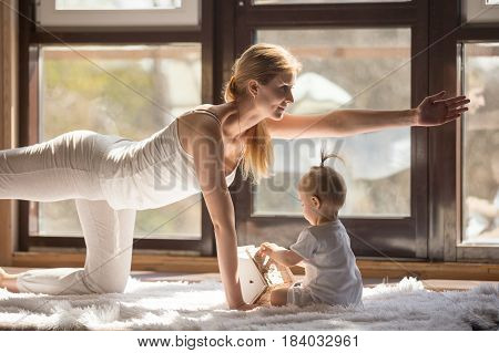 Young yogi smiling mother working out, doing Bird dog pose, wearing white sportswear, her baby daughter playing near her, yoga practice at home when having no time for gym. Healthy lifestyle concept