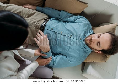 Overhead View Of Hypnosis Holding Wrist Of Bearded Man With Closed Eyes Lying On Couch