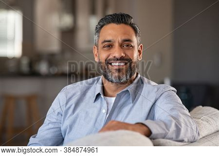 Portrait of happy mid adult man sitting on sofa at home. Handsome latin man in casual relaxing on couch and smiling. Cheerful indian guy looking at camera. 