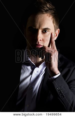 Low-key close up portrait of young serious businessman in dark suit sitting at desk looking straight, isolated on black background