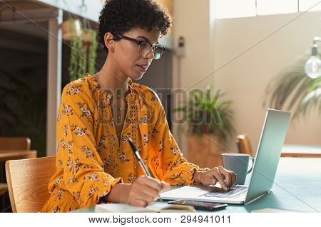 Focused college student sitting in cafeteria taking notes while using laptop. Young brazilian woman doing research for business at coffee shop. African american girl sitting in cafe writing notes.
