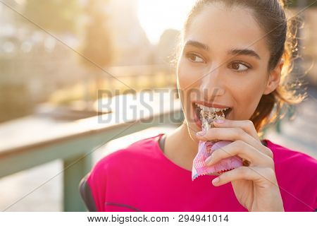 Athletic woman eating a protein bar. Closeup face of young sporty woman resting while biting a nutritive bar. Fitness beautiful woman eating a energy snack outdoor.