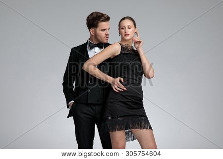 Confident groom standing behind his girfriend with his ahnd in his pocket while she is holding her hand on her hip and gesturing to wai on gray studio background