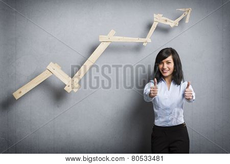 Concept: Succeeding in career or business. Young happy businesswoman holding thumbs up in front of positive trend graph,  isolated on grey background.