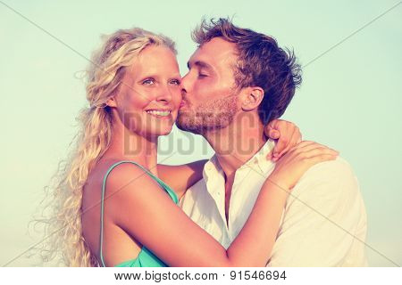 Romantic couple kissing in love enjoying sunset at beach. Young happy man kissing woman on cheek in romance on summer beach during honeymoon vacation holidays travel.