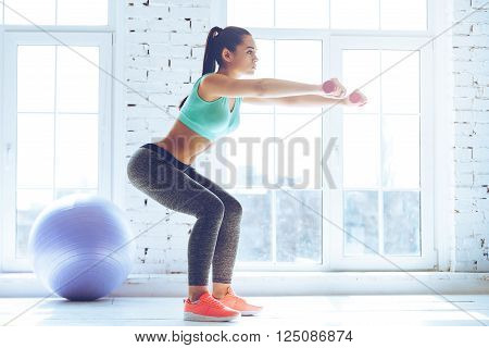 Deep squat. Side view of young beautiful woman in sportswear doing squat and holding dumbbells while standing in front of window at gym