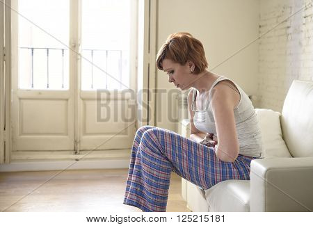 young beautiful red hair woman holding hurting tummy suffering stomach cramp and period pain sitting on home couch in painful face expression female menstruation concept