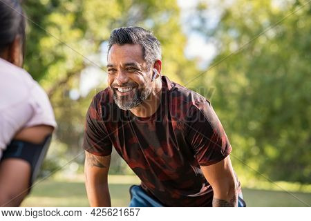 Happy mature fitness couple resting after a run in park. Smiling middle eastern man taking a break while looking at woman outdoor. Portrait of happy indian sportsman resting after jogging at park.