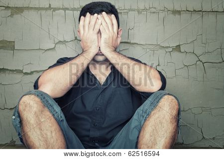 Depressed young man sitting on the floor and covering his face with a grunge wall background