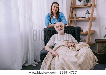 Smiling Female Nurse Standing Behind Senior Man In Chair