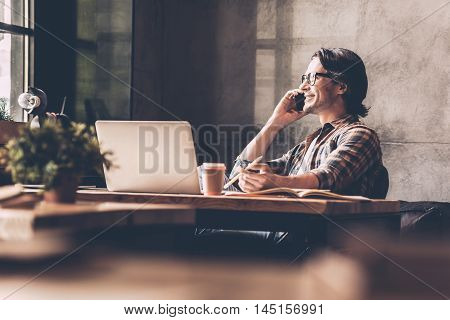 Staying in touch with his colleagues. Cheerful young man in casual wear looking through window and talking on the mobile phone while sitting near at the desk in creative office