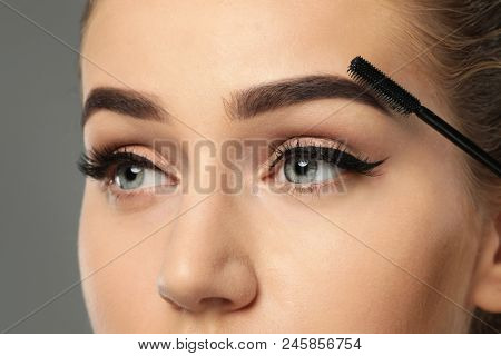 Young woman correcting shape of eyebrows on grey background, closeup