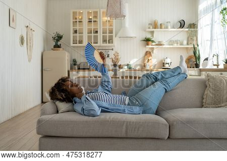 Unhappy Exhausted And Overheated Young African Woman Lying On Sofa At Home Waving Fan To Cool Down, 