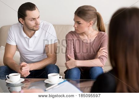 Young angry divorcing couple sitting on couch with arms crossed looking at each other with unconcealed hatred and hate, psychological help with problems in unhappy marriage, family counseling