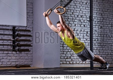 Young athlete man working out in gym pull ups with gymnastic rings against brick wall in the cross fit gym. Sportsman in the sportwear, t-shirt, shorts and sportive shoes. Concept of the cross fit activity.